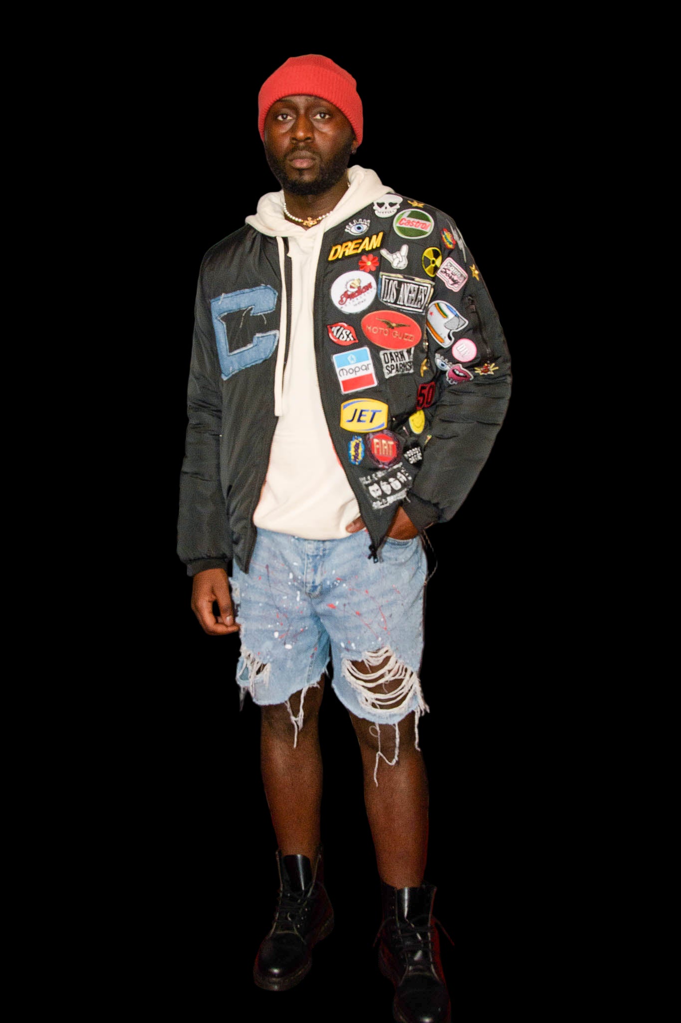 A person standing at a event wearing ripped denim shorts with red paint splatters on the leg, a hoodie with a logo, and a red beret.